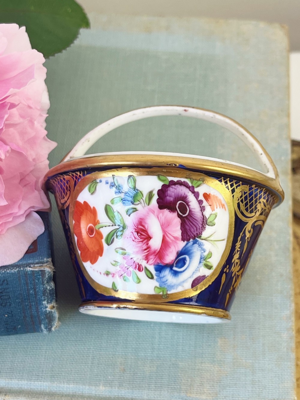 A miniature Regency Coalport porcelain basket featuring an overhead gilt handle, cobalt blue exterior casing with a hand-painted floral reserve, and a plain white interior. View II