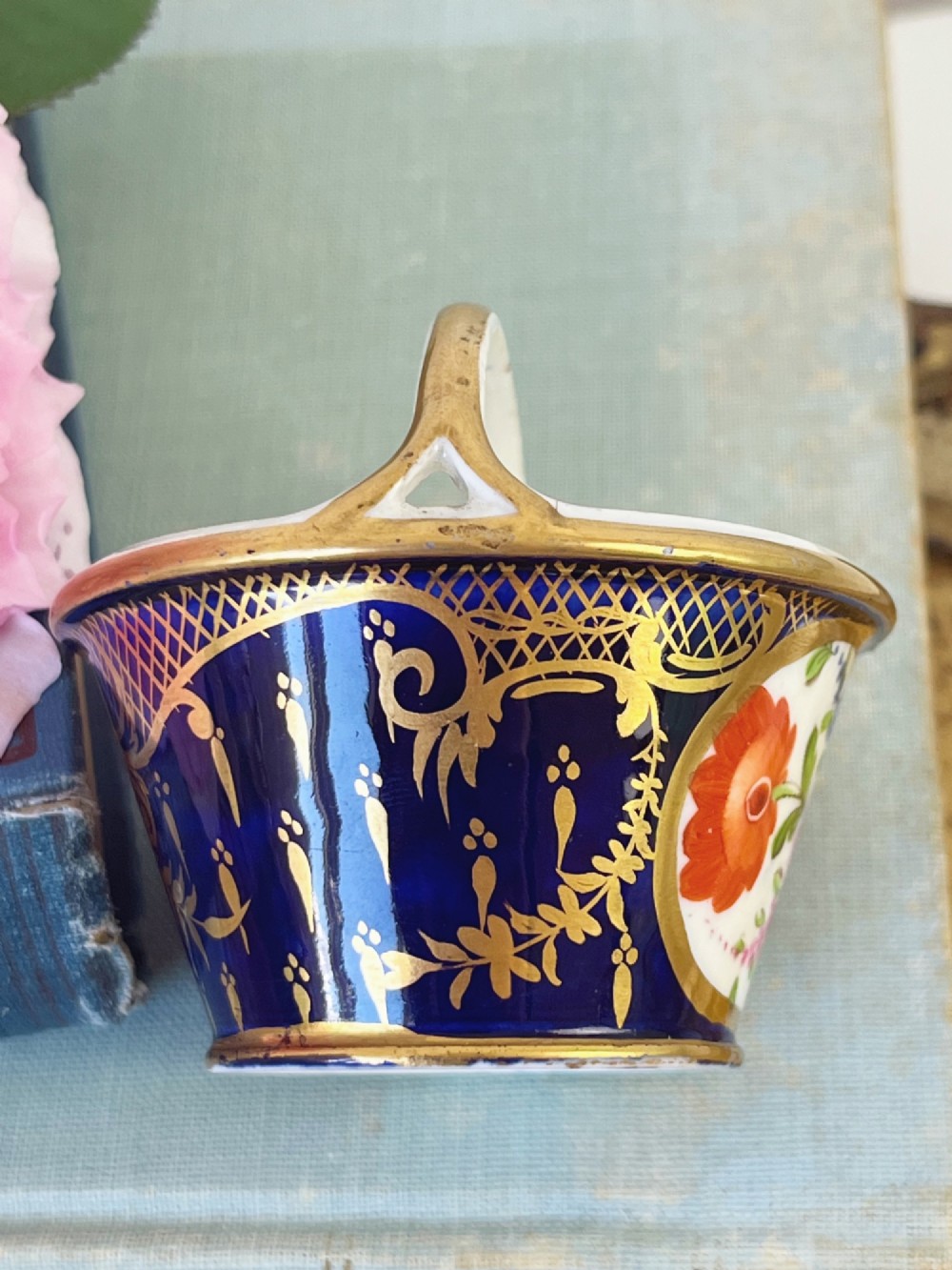 A miniature Regency Coalport porcelain basket featuring an overhead gilt handle, cobalt blue exterior casing with a hand-painted floral reserve, and a plain white interior. View V