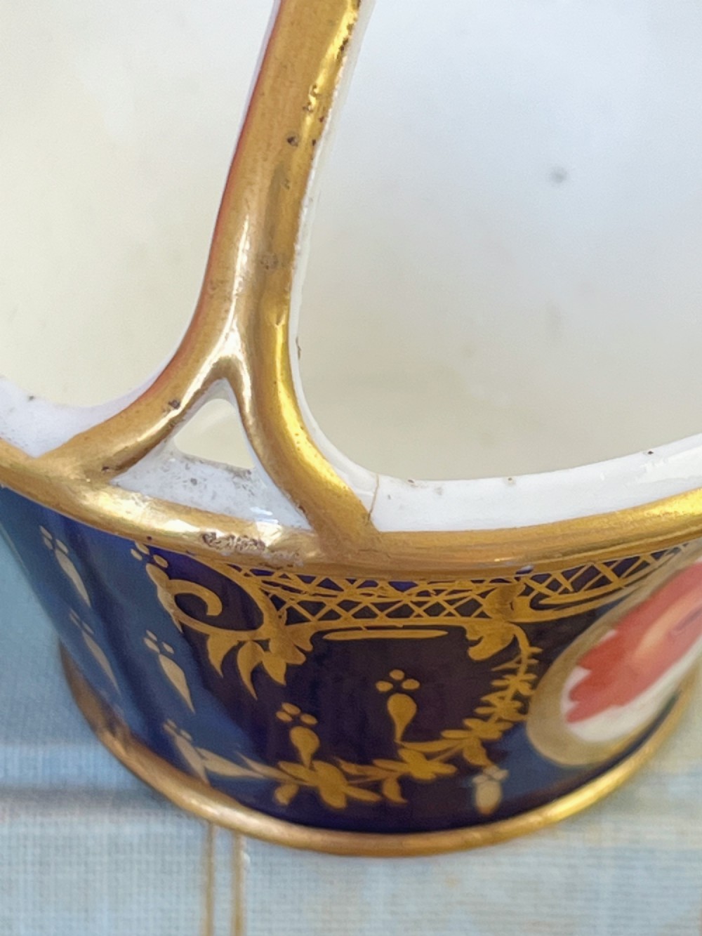 A miniature Regency Coalport porcelain basket featuring an overhead gilt handle, cobalt blue exterior casing with a hand-painted floral reserve, and a plain white interior. View VII