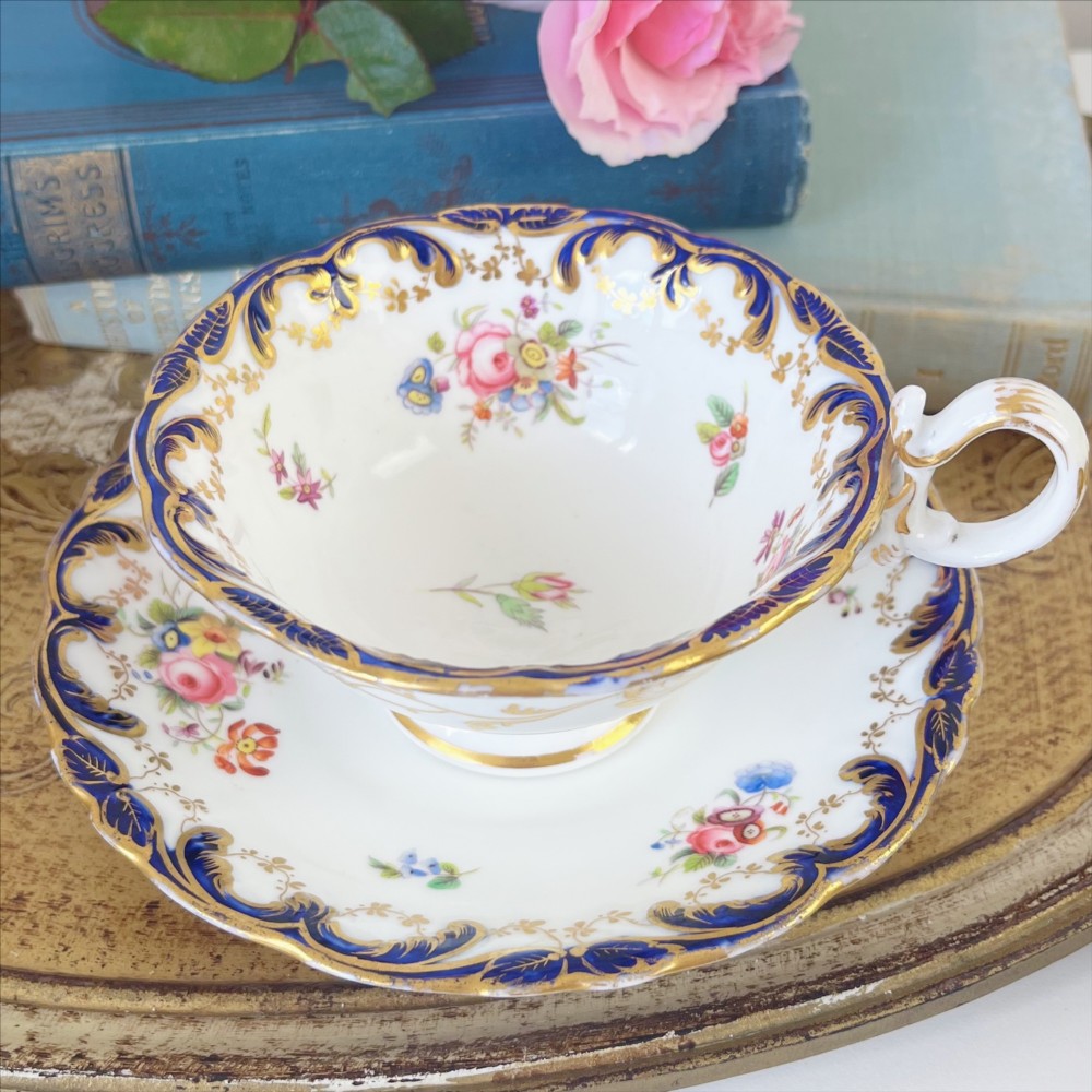 An antique bone china teacup and saucer set featuring cobalt blue scroll borders, gilded ornamentation, and hand-painted floral sprays on a white ground.
