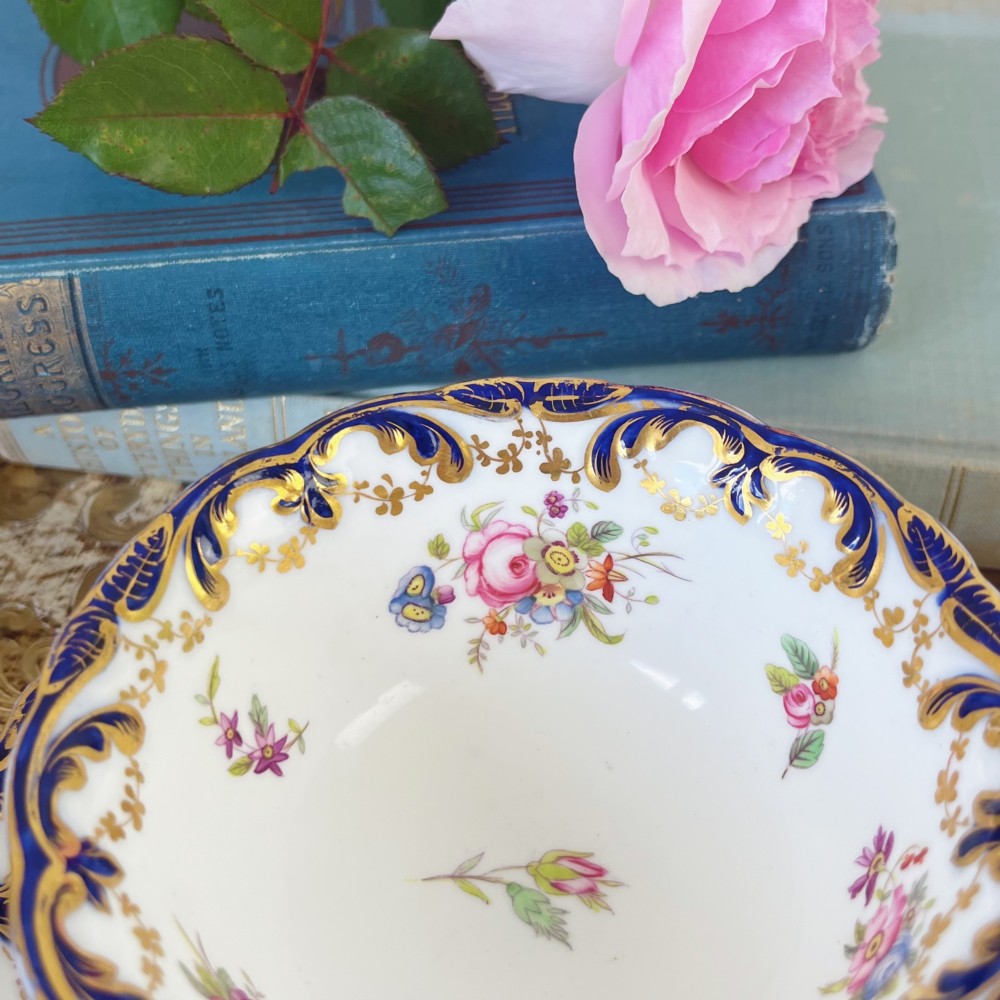 An antique bone china teacup and saucer set featuring cobalt blue scroll borders, gilded ornamentation, and hand-painted floral sprays on a white ground. View II
