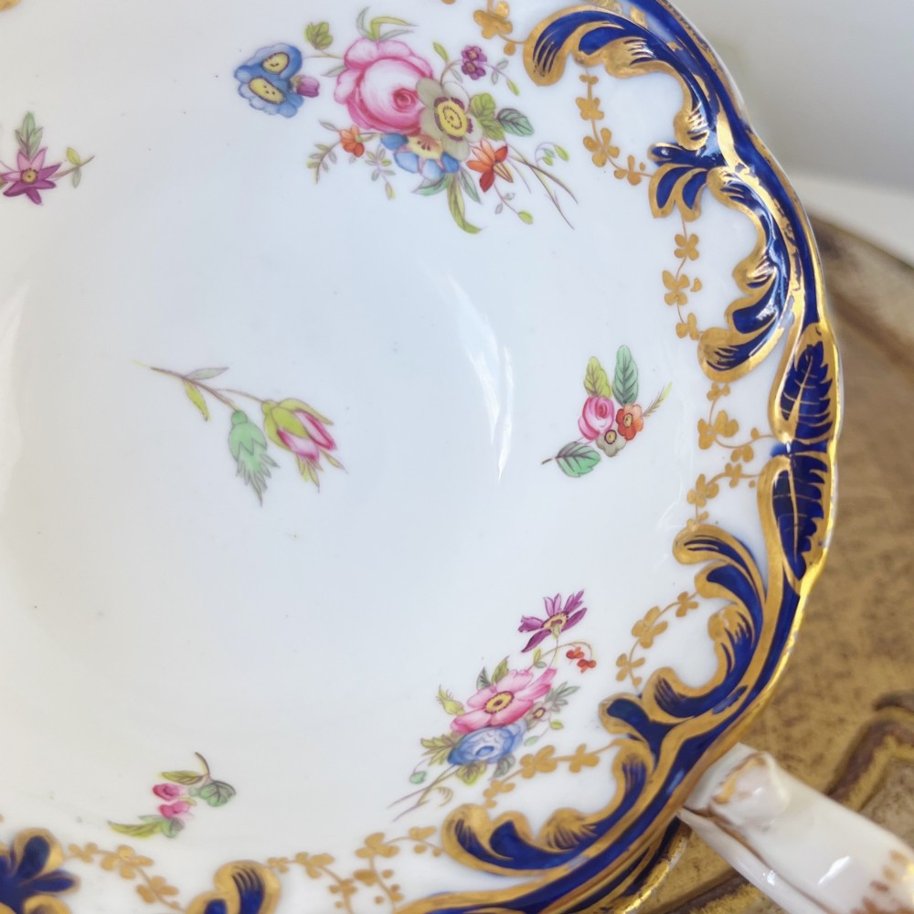 An antique bone china teacup and saucer set featuring cobalt blue scroll borders, gilded ornamentation, and hand-painted floral sprays on a white ground. View VIII