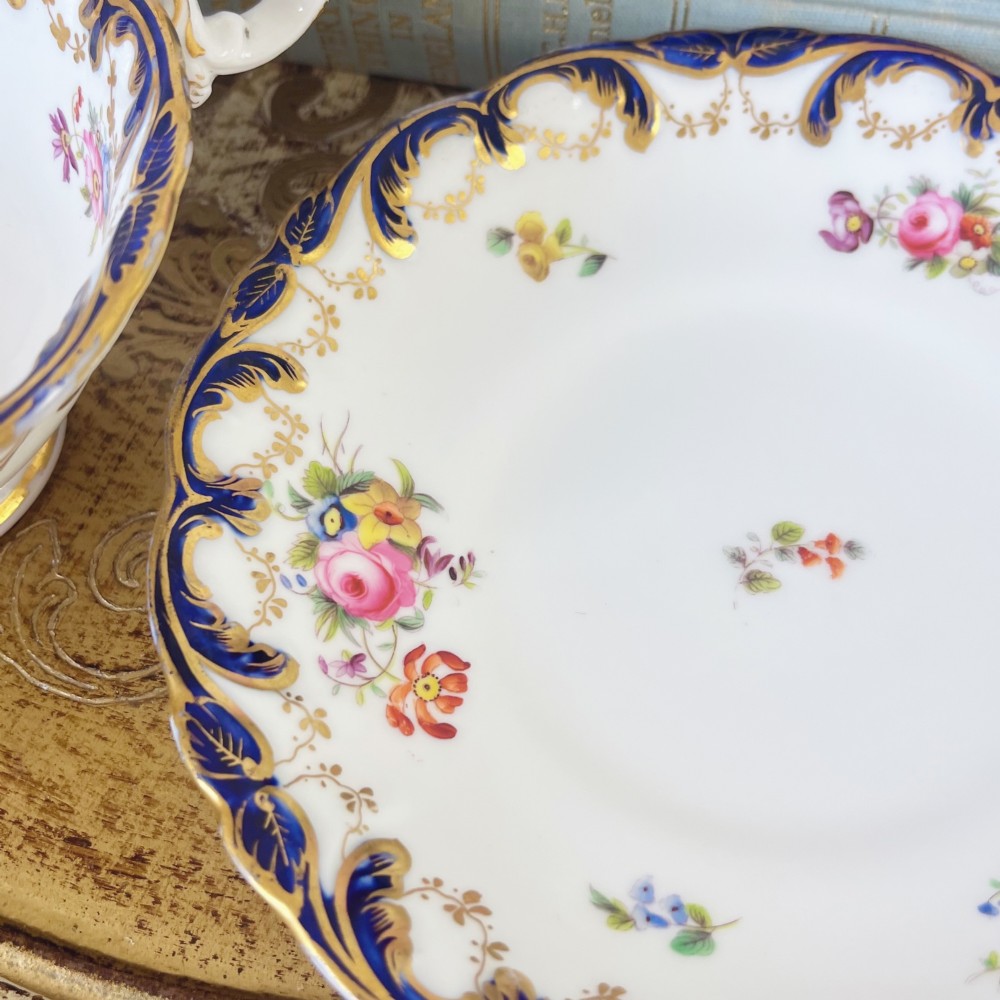 An antique bone china teacup and saucer set featuring cobalt blue scroll borders, gilded ornamentation, and hand-painted floral sprays on a white ground. View X