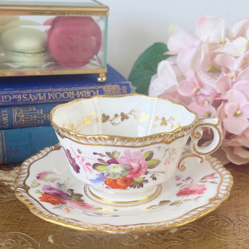 A white porcelain teacup and saucer set featuring hand-painted floral motifs, a gadrooned rim, and gilded scroll accents.