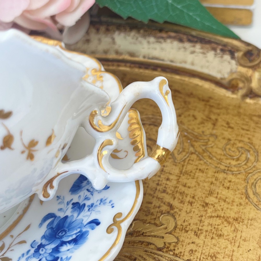 An antique Regency period white porcelain teacup and saucer featuring hand-painted blue floral motifs and gilded ornate rim embellishments. View II
