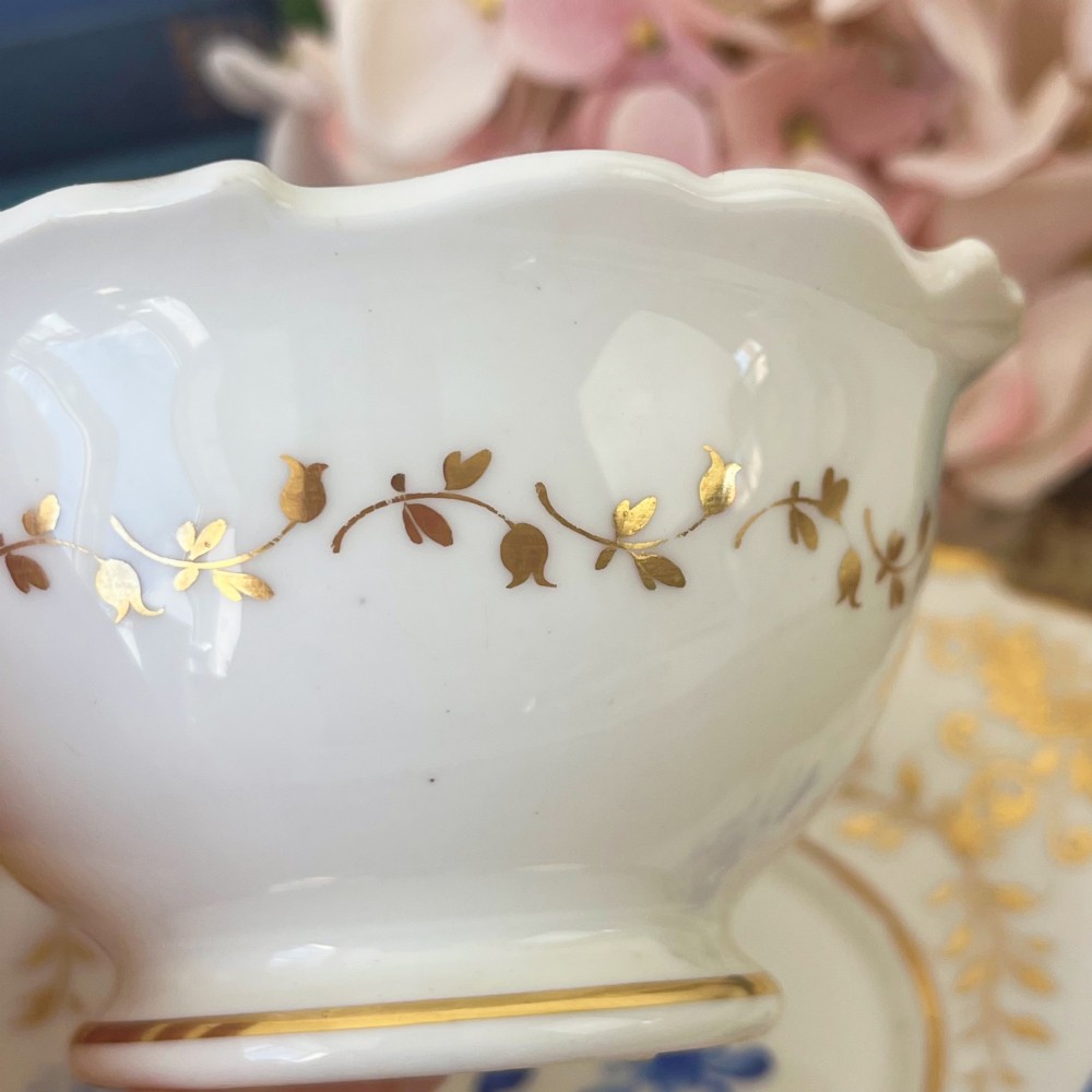 An antique Regency period white porcelain teacup and saucer featuring hand-painted blue floral motifs and gilded ornate rim embellishments. View IV