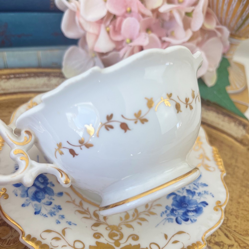 An antique Regency period white porcelain teacup and saucer featuring hand-painted blue floral motifs and gilded ornate rim embellishments. View V