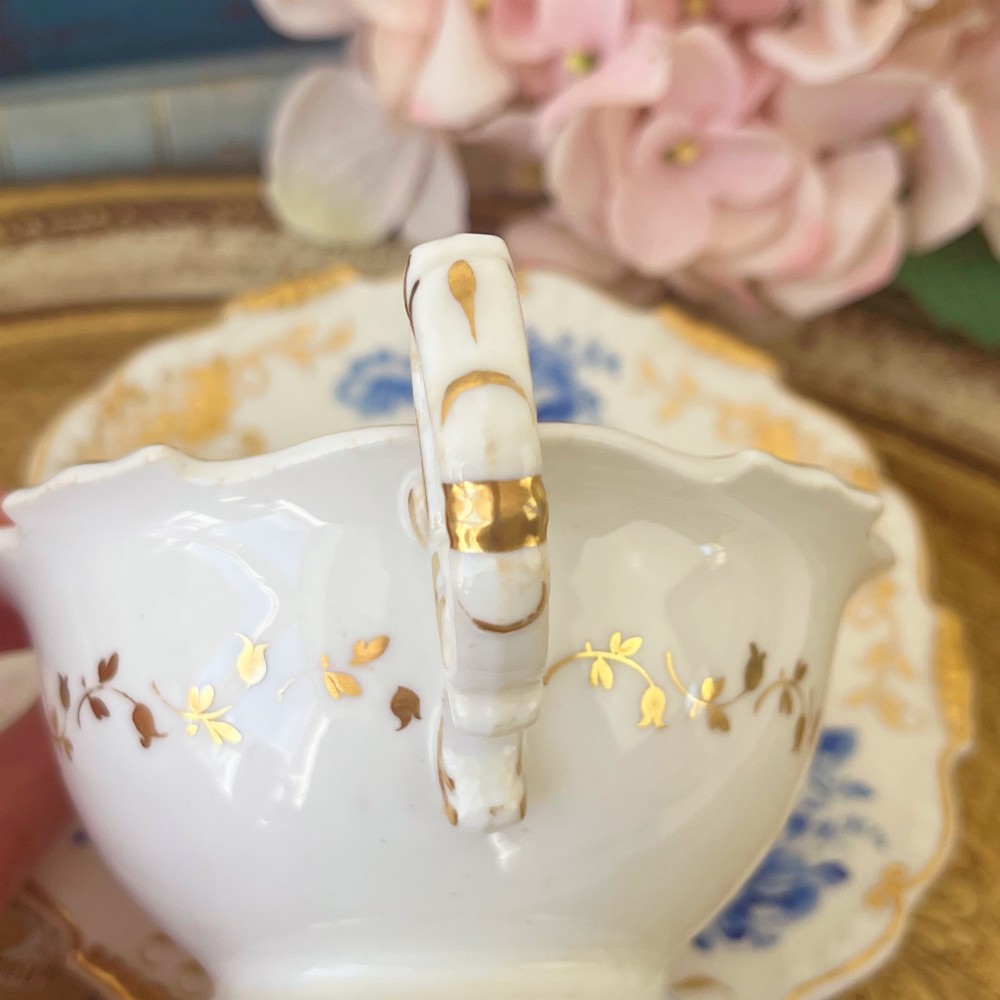 An antique Regency period white porcelain teacup and saucer featuring hand-painted blue floral motifs and gilded ornate rim embellishments. View VI