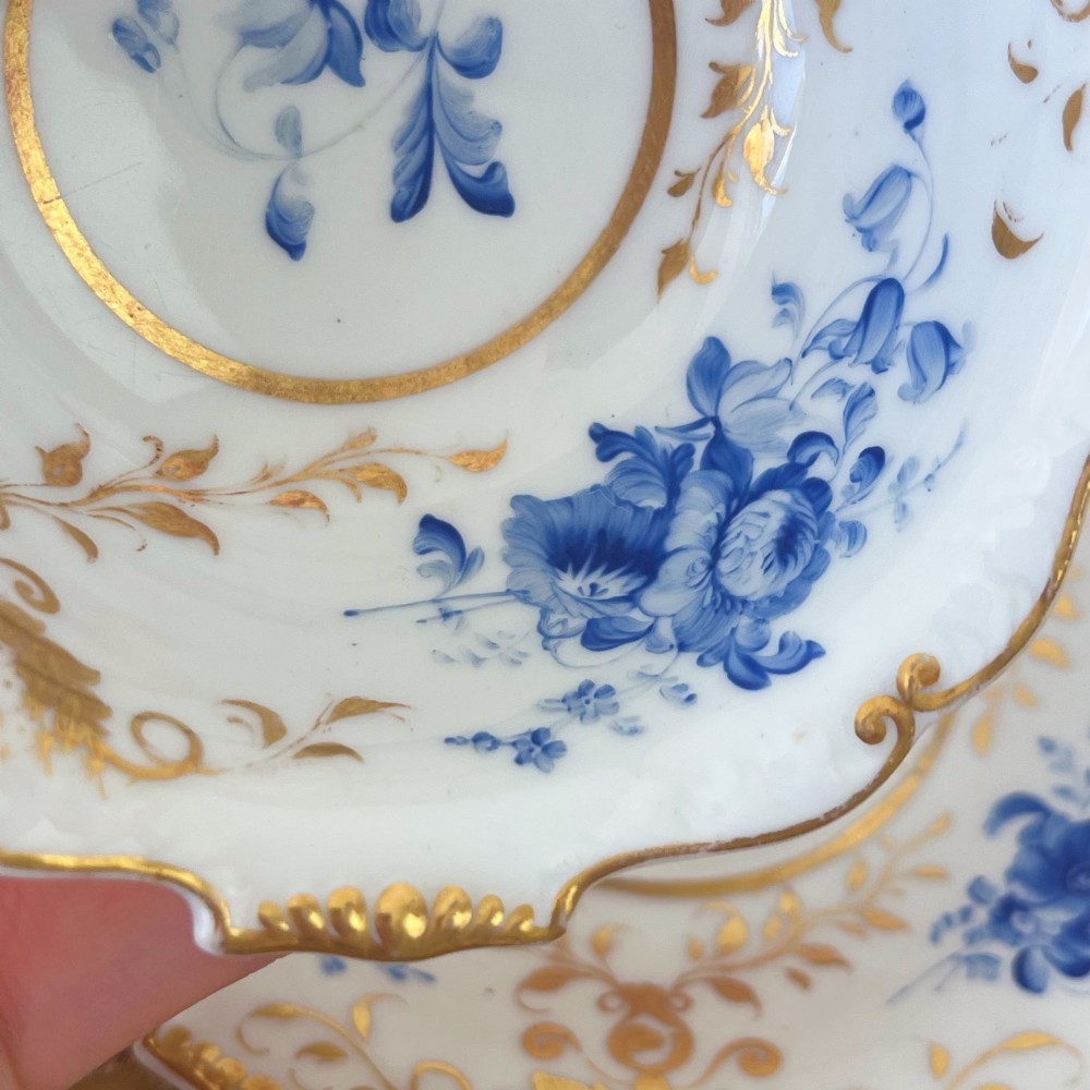 An antique Regency period white porcelain teacup and saucer featuring hand-painted blue floral motifs and gilded ornate rim embellishments. View VIII