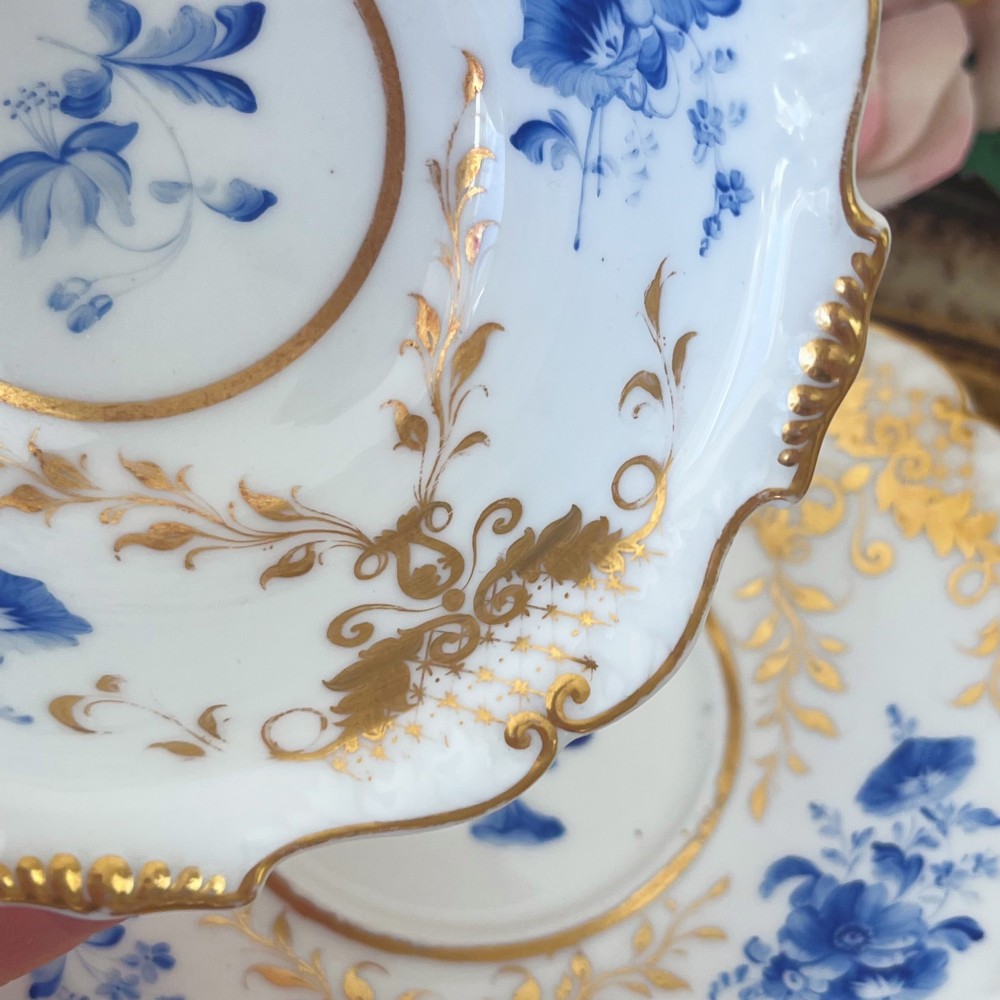 An antique Regency period white porcelain teacup and saucer featuring hand-painted blue floral motifs and gilded ornate rim embellishments. View IX