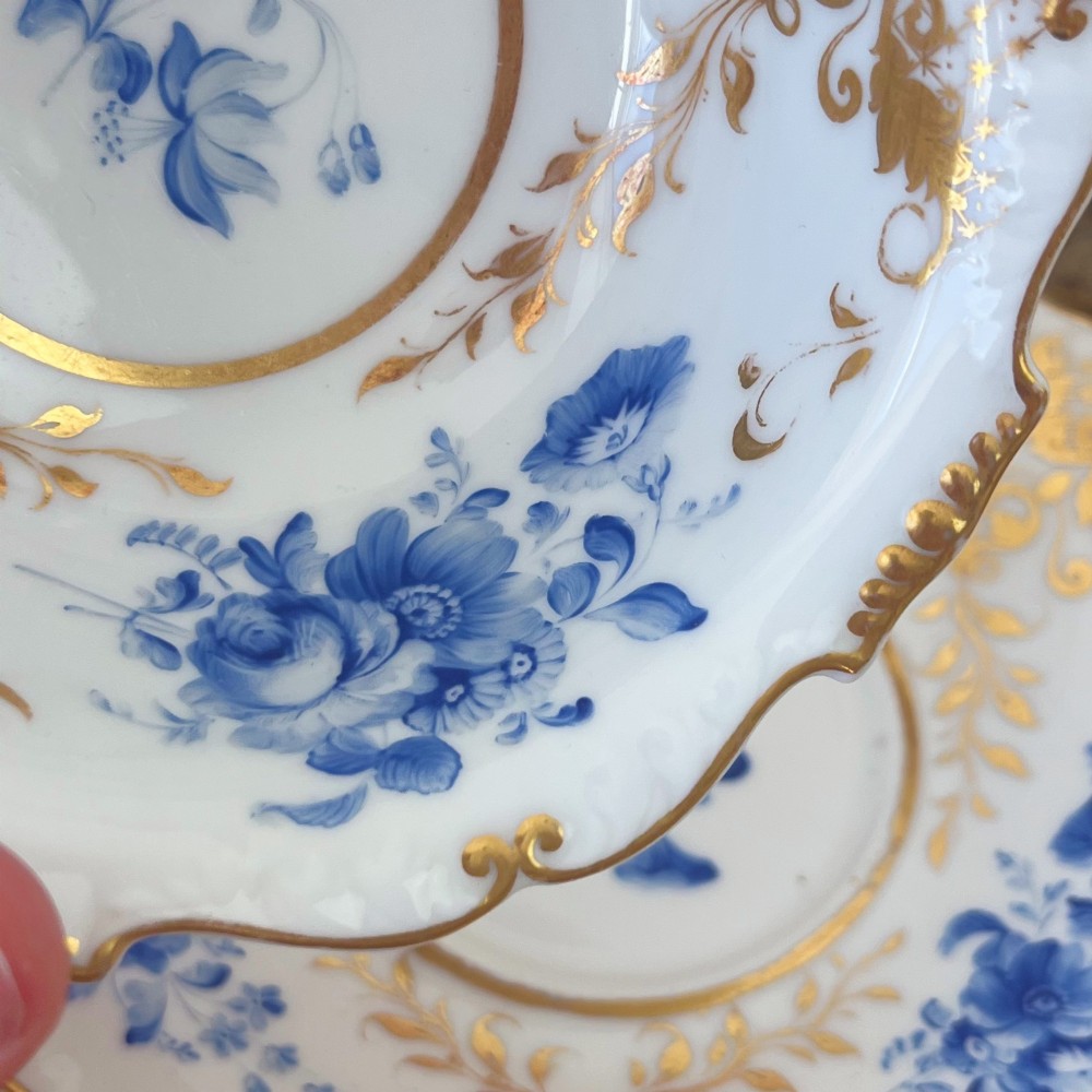 An antique Regency period white porcelain teacup and saucer featuring hand-painted blue floral motifs and gilded ornate rim embellishments. View X