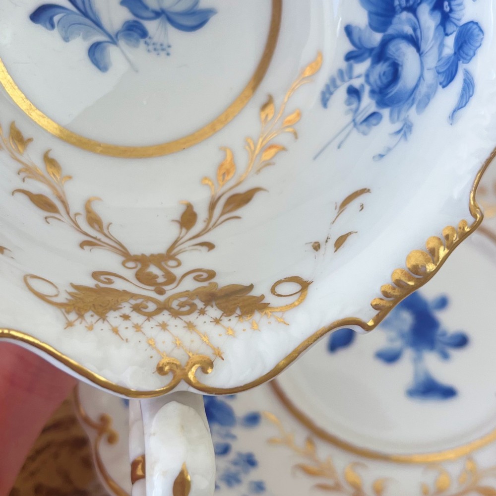 An antique Regency period white porcelain teacup and saucer featuring hand-painted blue floral motifs and gilded ornate rim embellishments. View XI