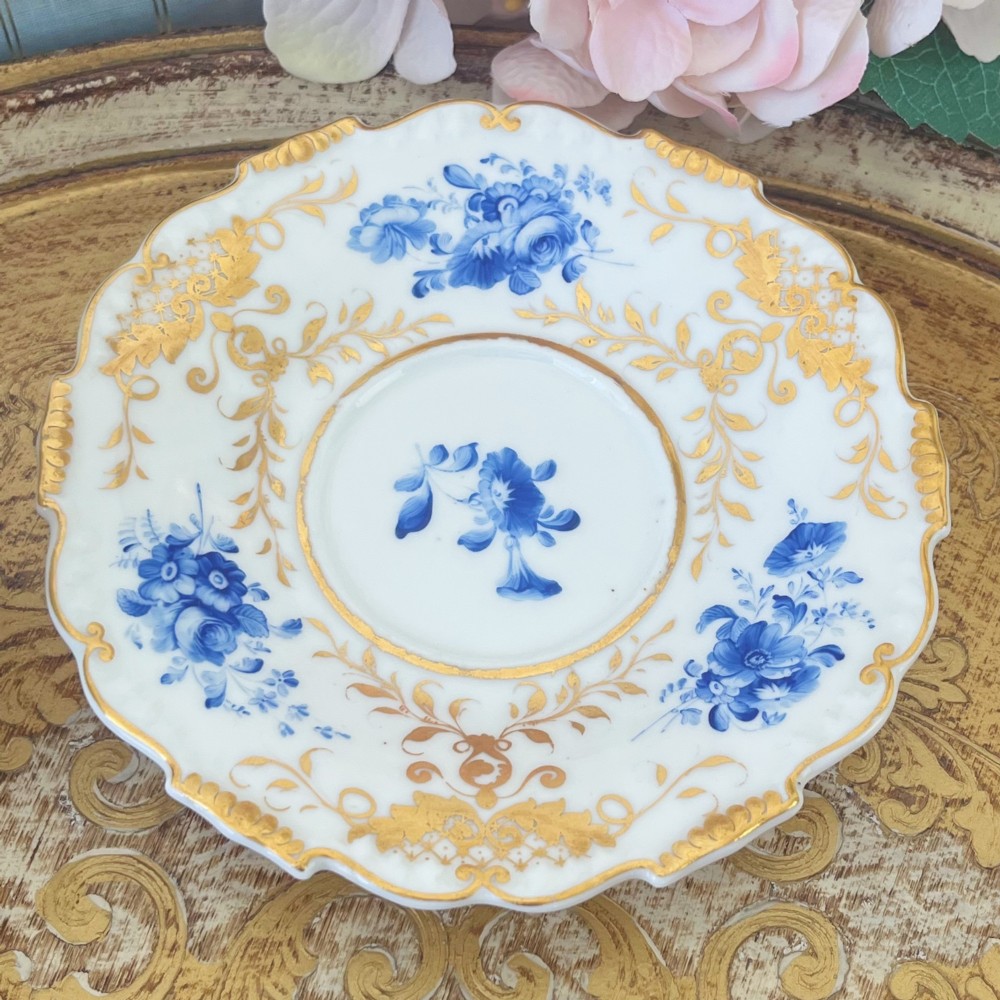 An antique Regency period white porcelain teacup and saucer featuring hand-painted blue floral motifs and gilded ornate rim embellishments. View XII