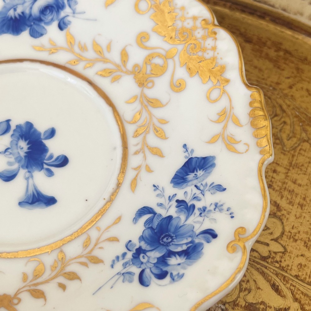 An antique Regency period white porcelain teacup and saucer featuring hand-painted blue floral motifs and gilded ornate rim embellishments. View XIII
