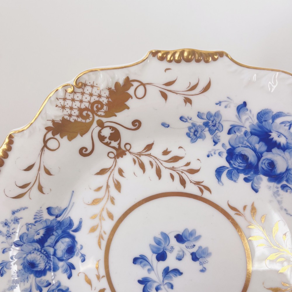 An early nineteenth-century white porcelain plate featuring hand-painted blue floral clusters, ornate gilded foliage along the scalloped rim, and a central circular gilt border. View III