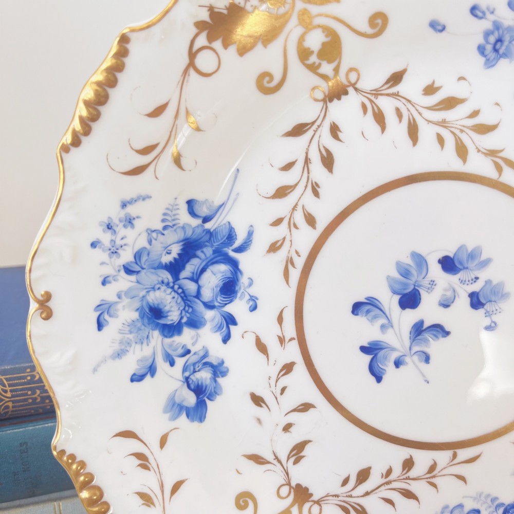 An early nineteenth-century white porcelain plate featuring hand-painted blue floral clusters, ornate gilded foliage along the scalloped rim, and a central circular gilt border. View IV
