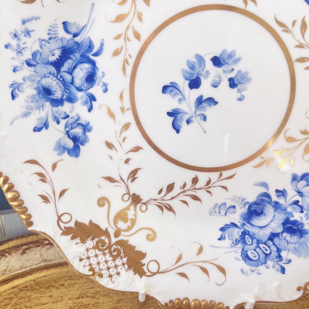 An early nineteenth-century white porcelain plate featuring hand-painted blue floral clusters, ornate gilded foliage along the scalloped rim, and a central circular gilt border. View V