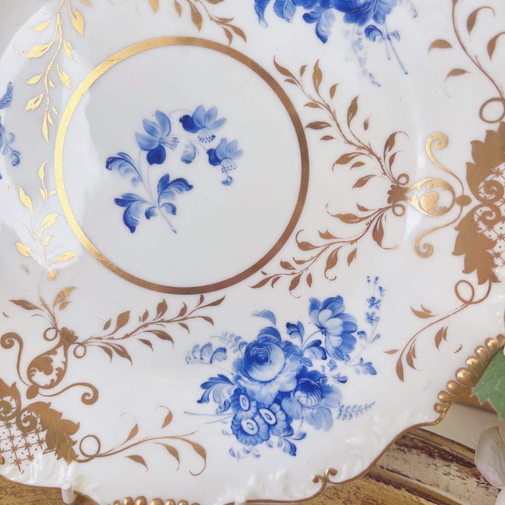 An early nineteenth-century white porcelain plate featuring hand-painted blue floral clusters, ornate gilded foliage along the scalloped rim, and a central circular gilt border. View VI