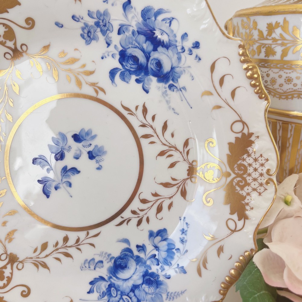 An early nineteenth-century white porcelain plate featuring hand-painted blue floral clusters, ornate gilded foliage along the scalloped rim, and a central circular gilt border. View VII