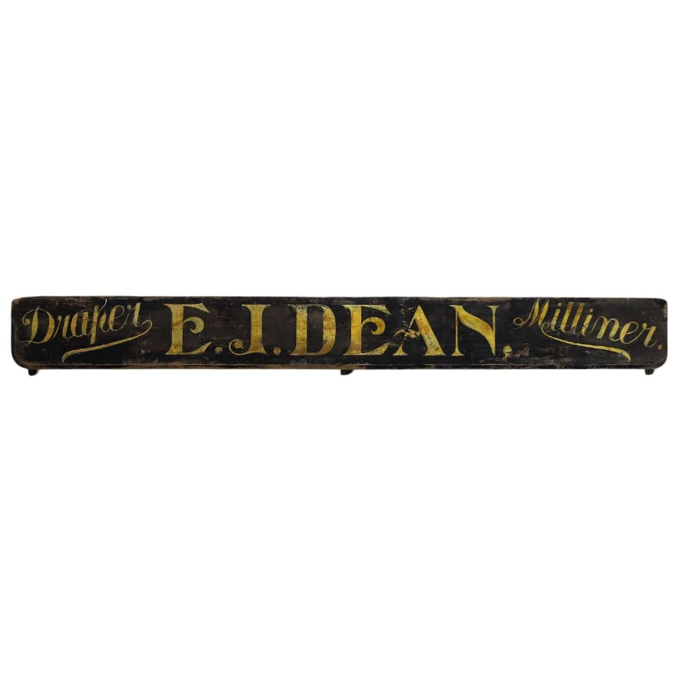 A long, dark rectangular pine shop sign featuring hand-painted gold lettering that reads Draper E.J. DEAN. Milliner.
