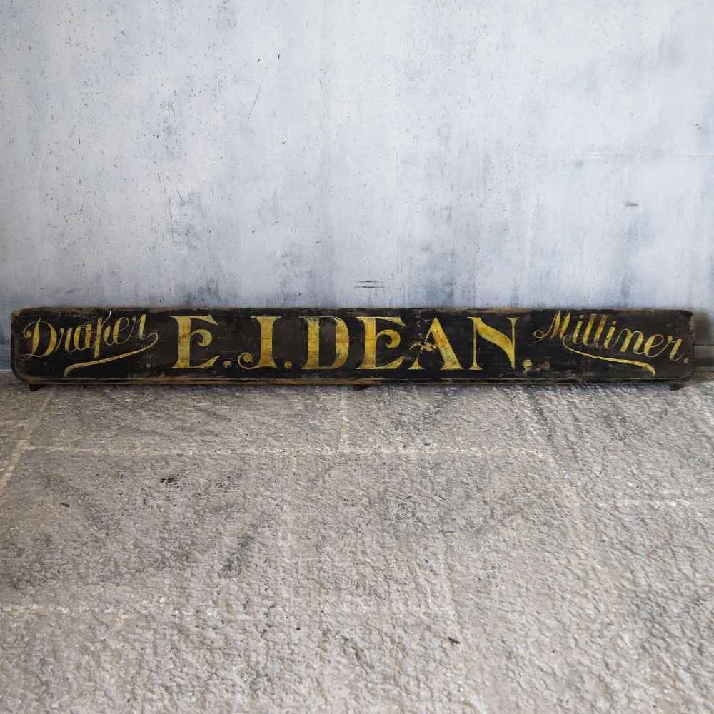 A long, dark rectangular pine shop sign featuring hand-painted gold lettering that reads Draper E.J. DEAN. Milliner. View II
