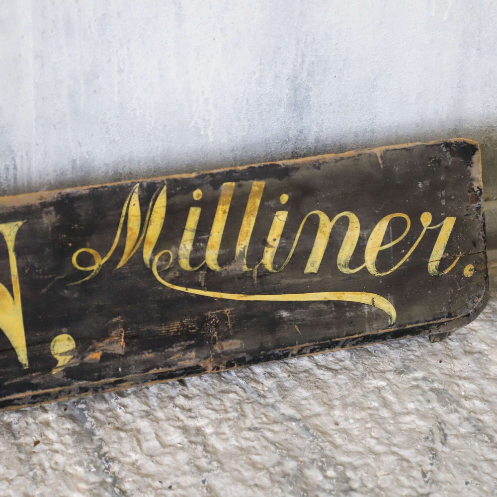 A long, dark rectangular pine shop sign featuring hand-painted gold lettering that reads Draper E.J. DEAN. Milliner. View III