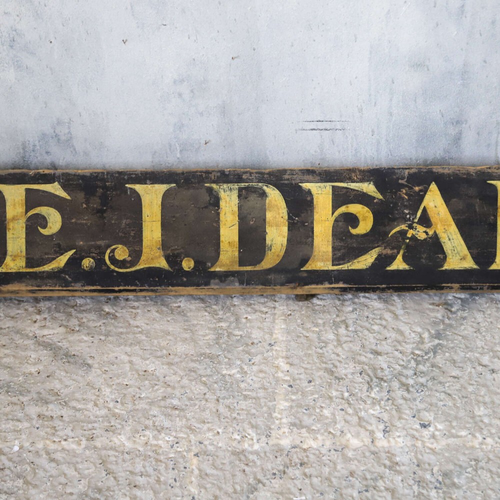 A long, dark rectangular pine shop sign featuring hand-painted gold lettering that reads Draper E.J. DEAN. Milliner. View IV