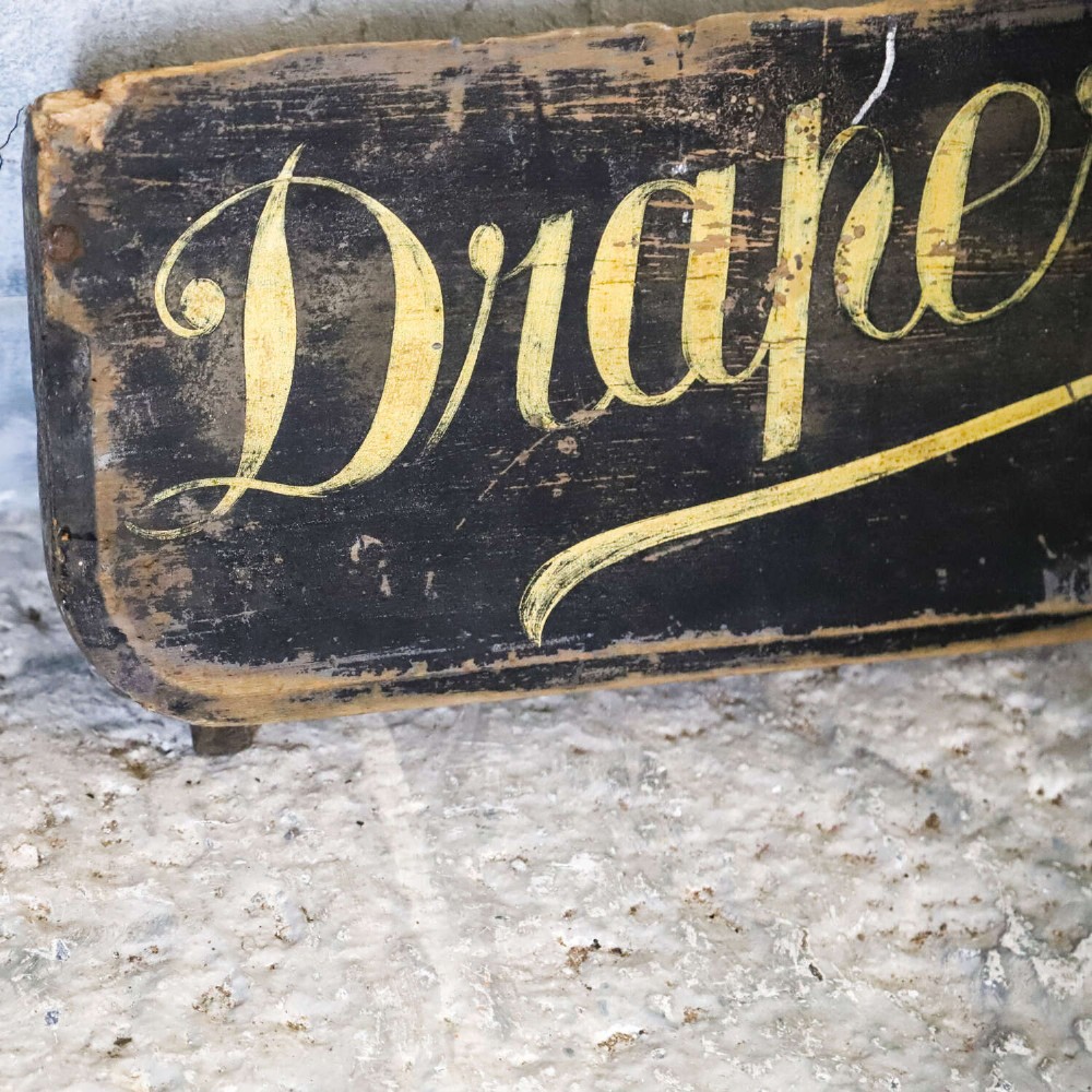 A long, dark rectangular pine shop sign featuring hand-painted gold lettering that reads Draper E.J. DEAN. Milliner. View V