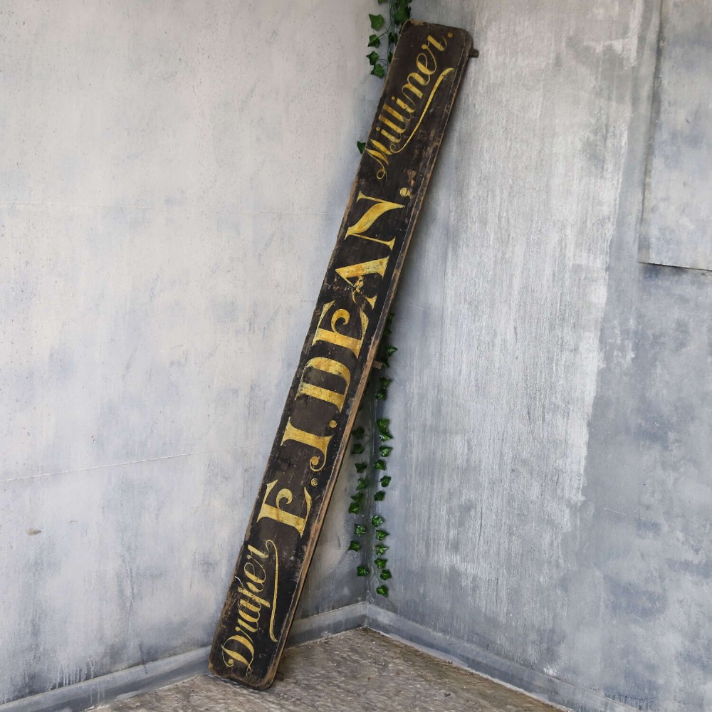 A long, dark rectangular pine shop sign featuring hand-painted gold lettering that reads Draper E.J. DEAN. Milliner. View VII