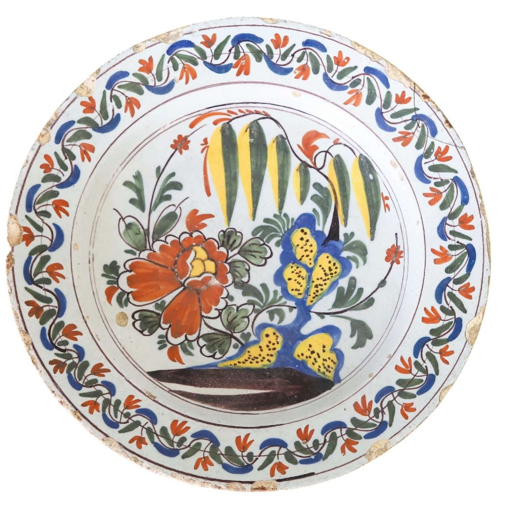 A circular 18th-century English Delftware charger featuring hand-painted polychrome floral motifs, rockwork, and a scrolling border on an off-white background.