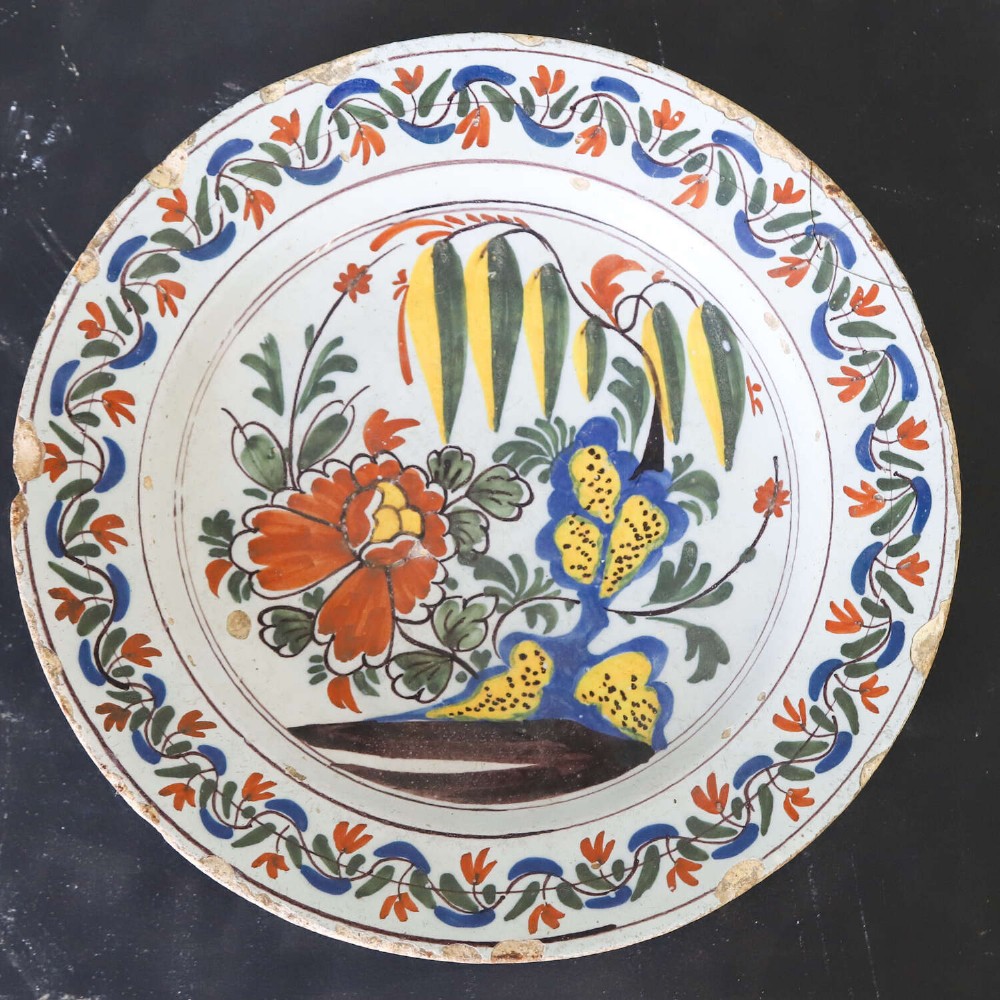 A circular 18th-century English Delftware charger featuring hand-painted polychrome floral motifs, rockwork, and a scrolling border on an off-white background. View II