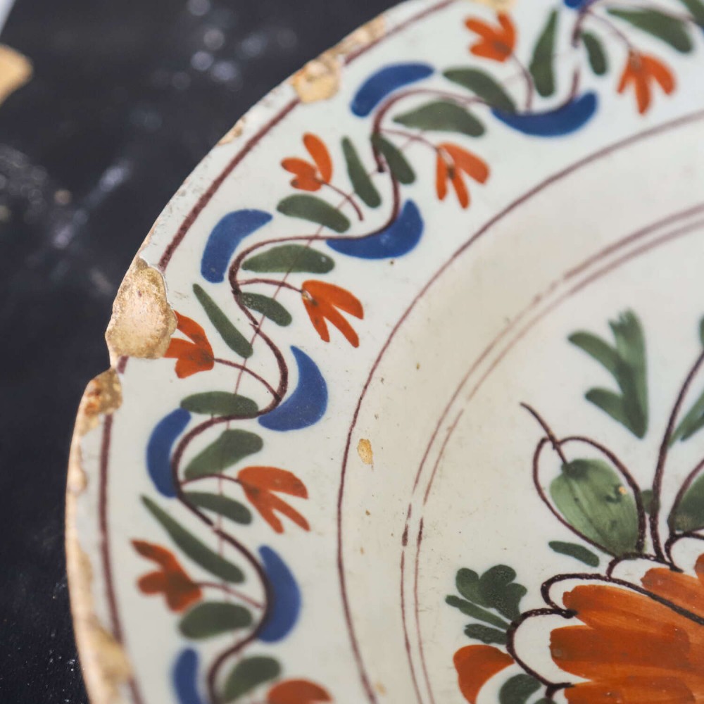 A circular 18th-century English Delftware charger featuring hand-painted polychrome floral motifs, rockwork, and a scrolling border on an off-white background. View III
