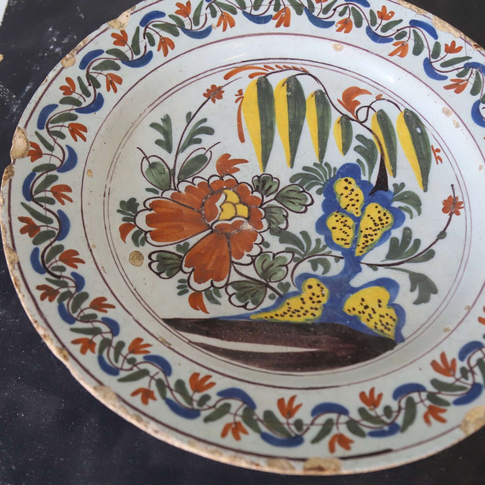 A circular 18th-century English Delftware charger featuring hand-painted polychrome floral motifs, rockwork, and a scrolling border on an off-white background. View IV