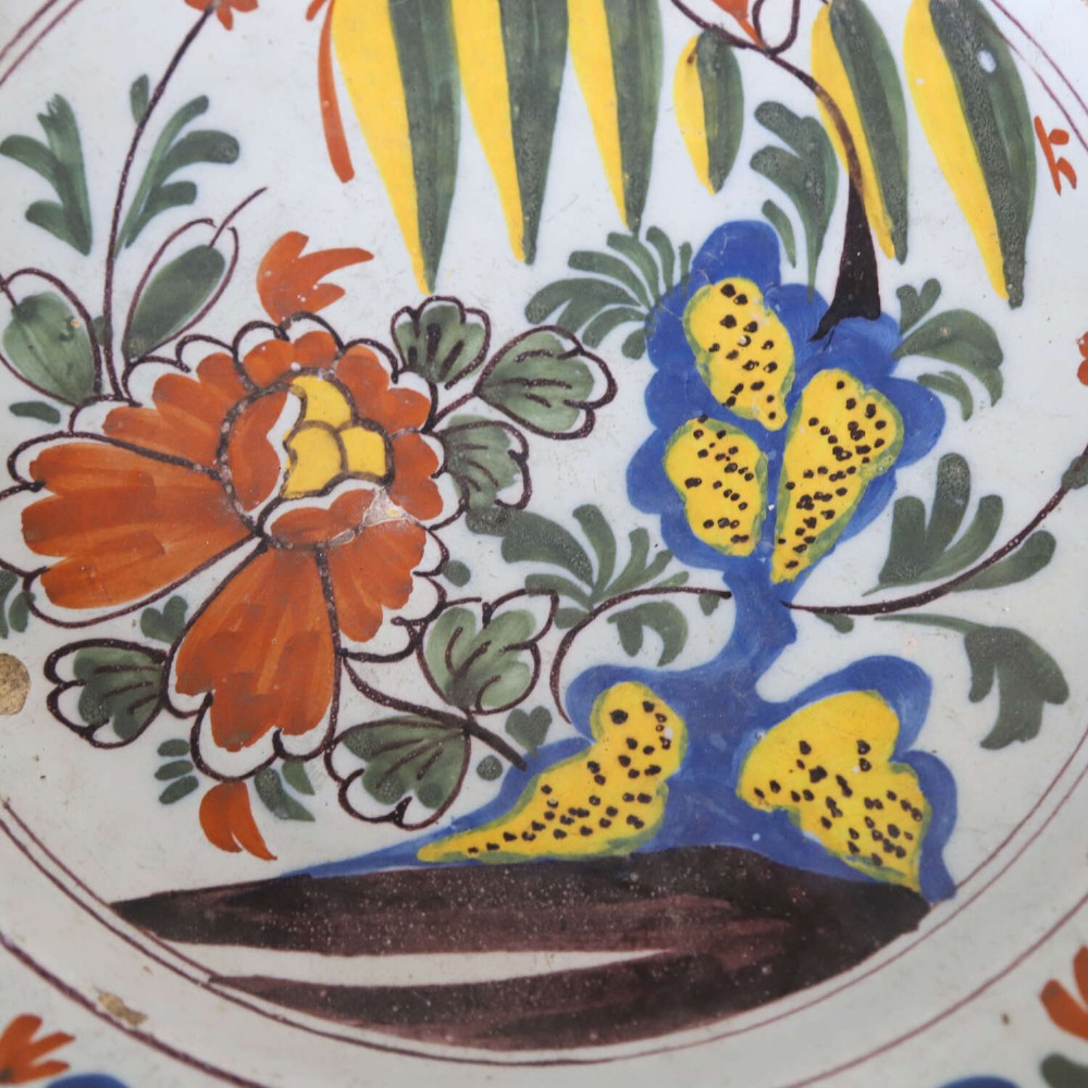 A circular 18th-century English Delftware charger featuring hand-painted polychrome floral motifs, rockwork, and a scrolling border on an off-white background. View V