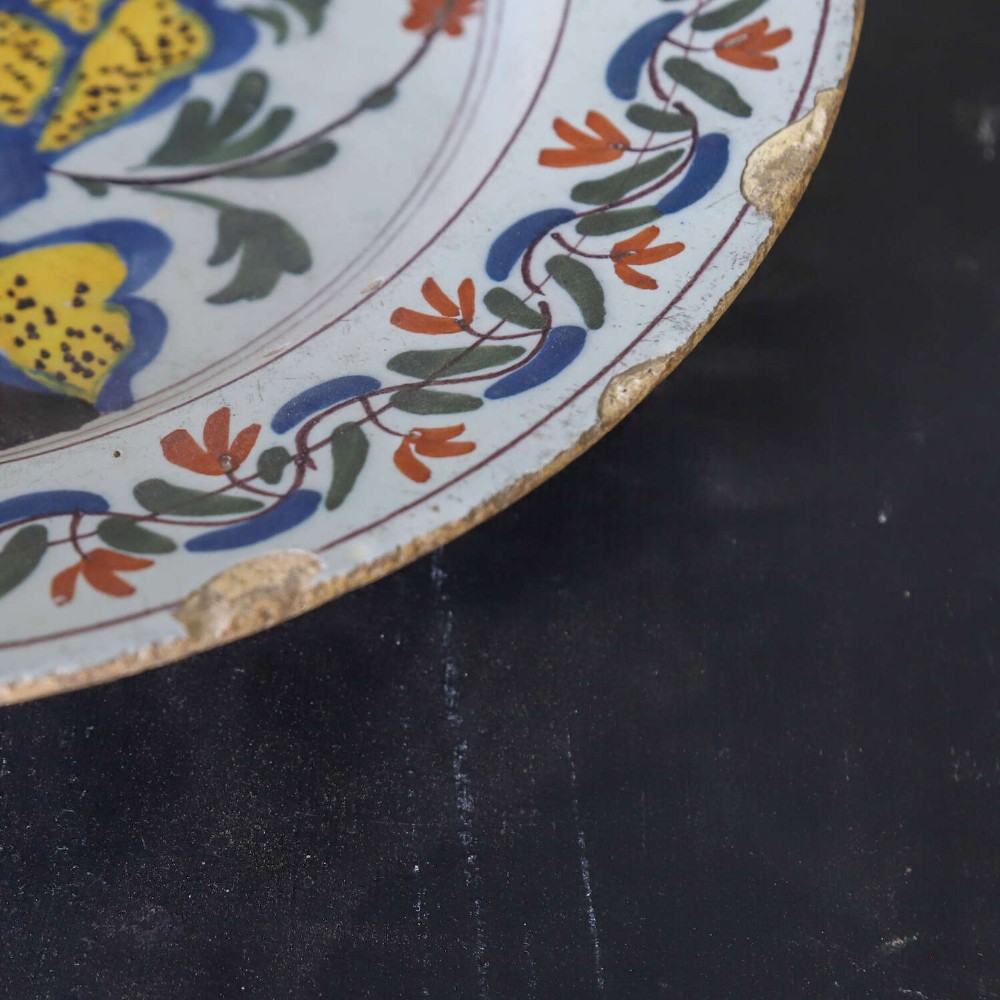 A circular 18th-century English Delftware charger featuring hand-painted polychrome floral motifs, rockwork, and a scrolling border on an off-white background. View VI