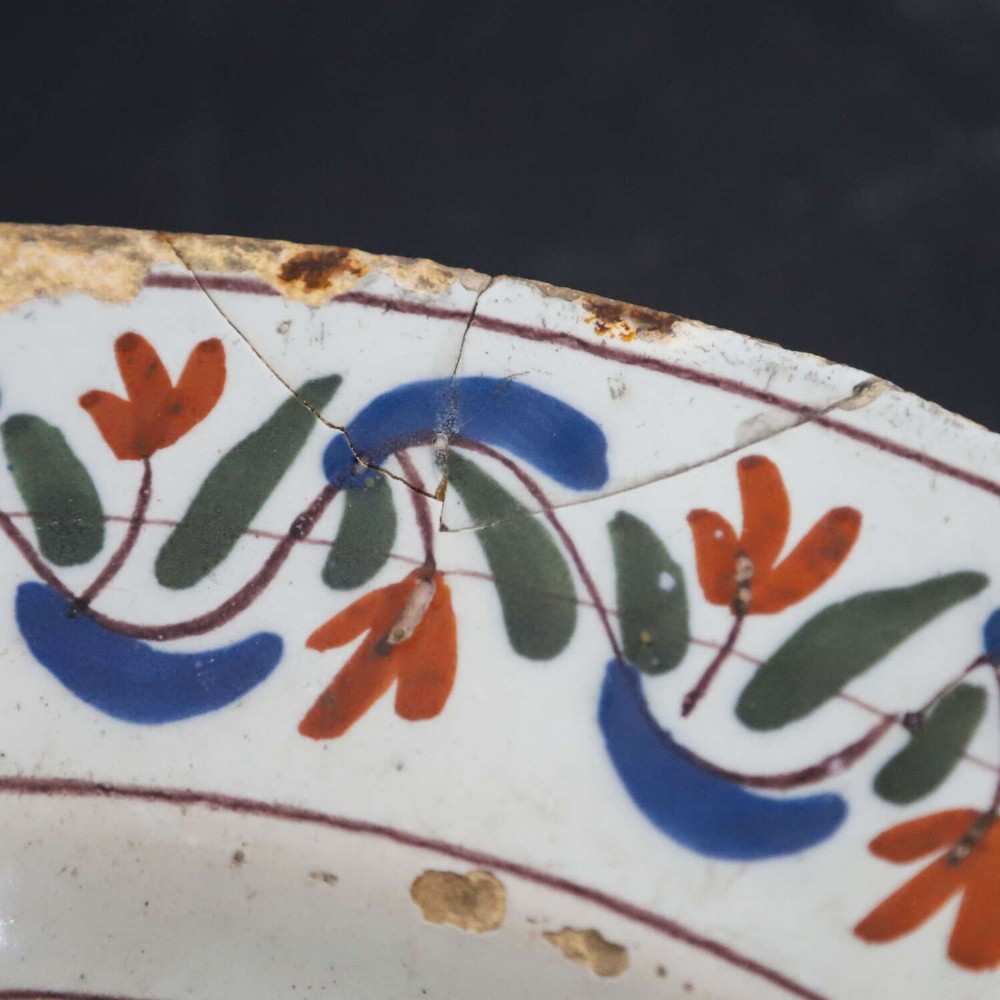 A circular 18th-century English Delftware charger featuring hand-painted polychrome floral motifs, rockwork, and a scrolling border on an off-white background. View VII