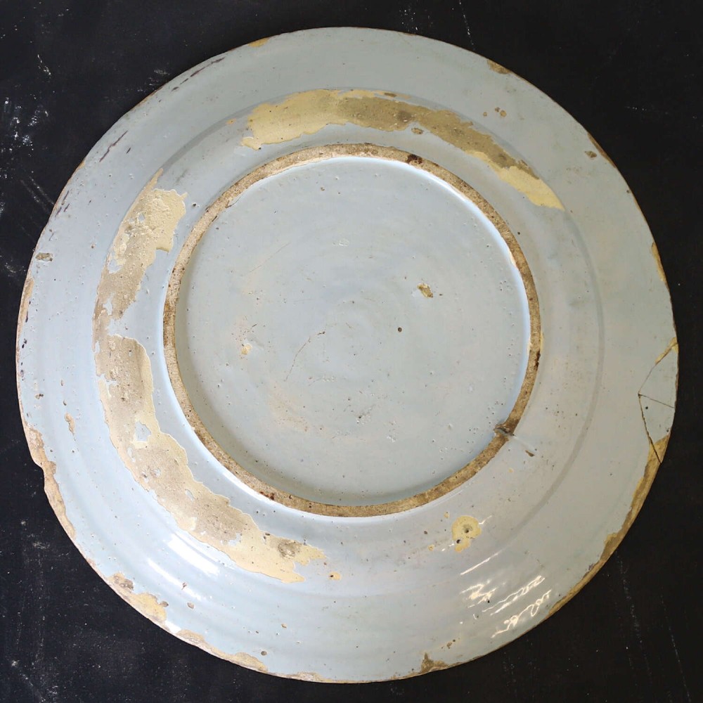 A circular 18th-century English Delftware charger featuring hand-painted polychrome floral motifs, rockwork, and a scrolling border on an off-white background. View IX