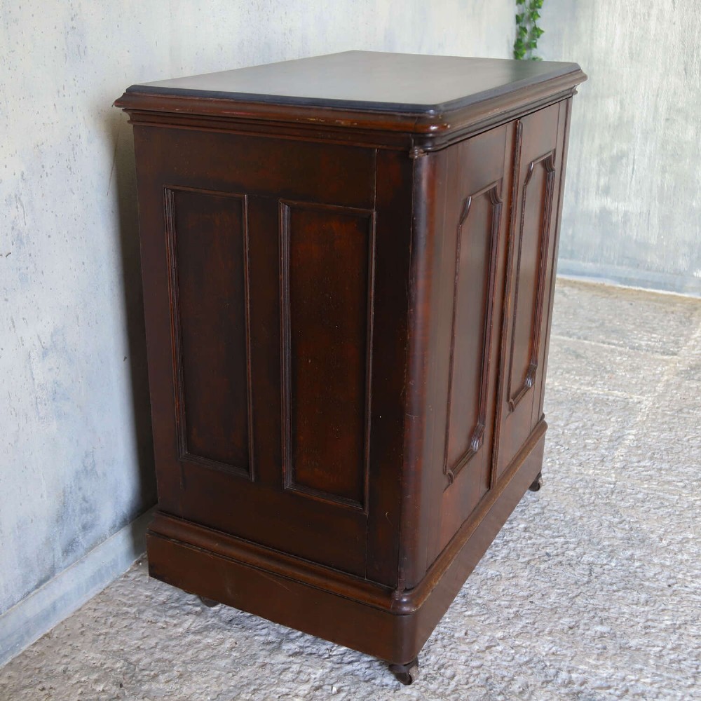 A late 19th-century dark mahogany office cupboard featuring two recessed panel doors, central brass keyhole escutcheon, plinth base, and fitted metal castors. View X