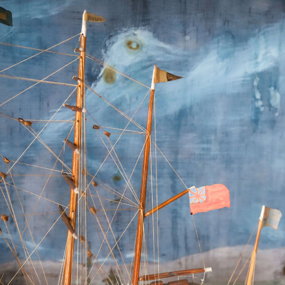 A glazed wooden shadow box containing a three-dimensional diorama of a four-masted sailing ship and a steam barge against a painted blue background. View IV