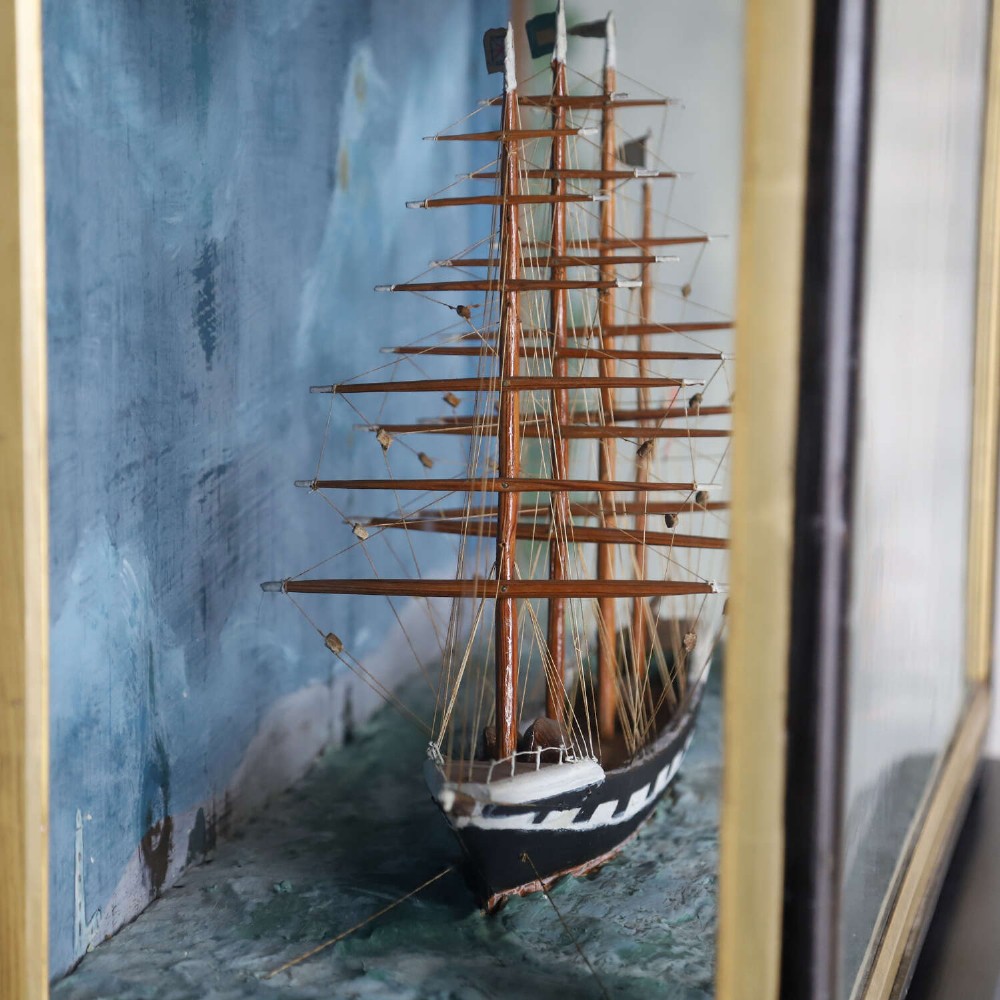 A glazed wooden shadow box containing a three-dimensional diorama of a four-masted sailing ship and a steam barge against a painted blue background. View VI