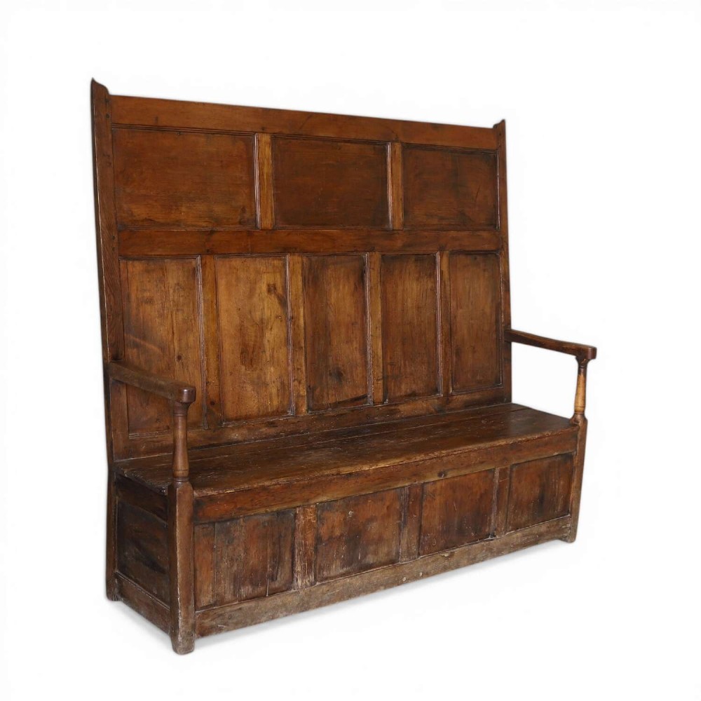 A high-backed antique wooden settle featuring raised timber panels, flat armrests with turned supports, and an enclosed panelled base.