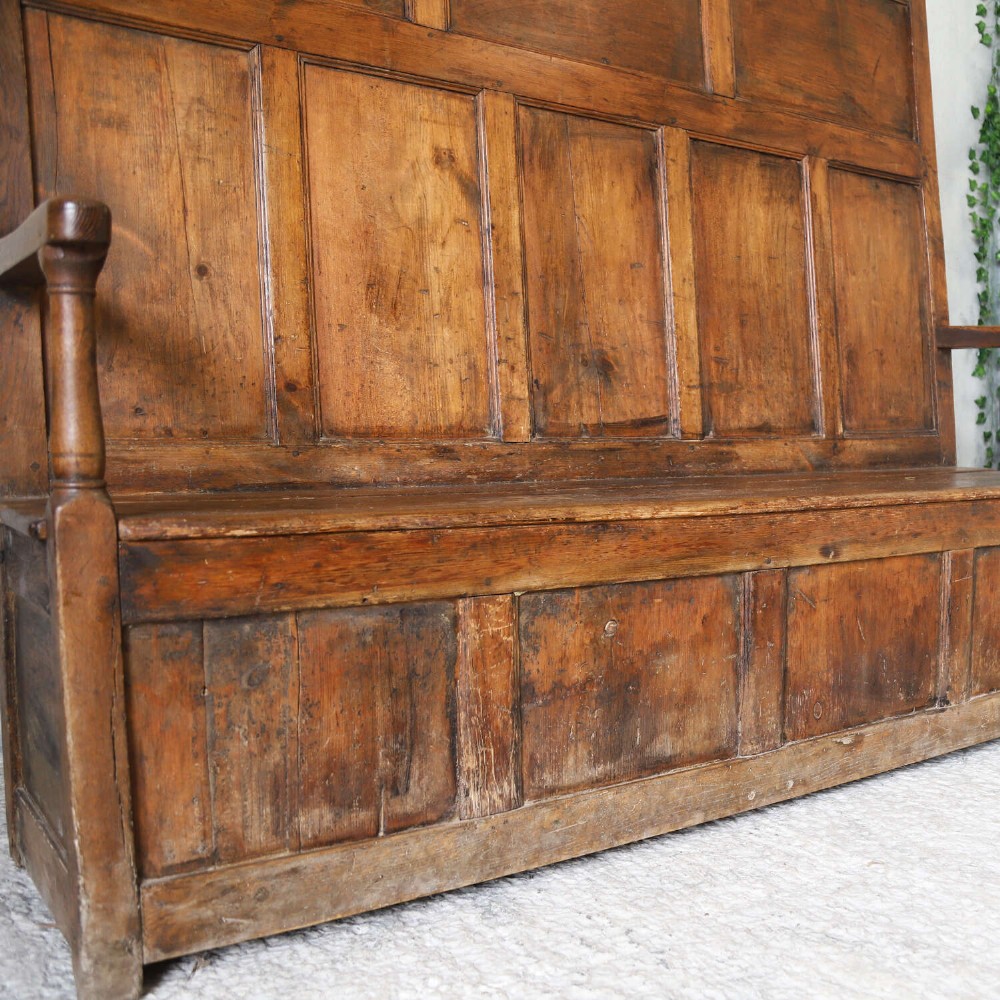 A high-backed antique wooden settle featuring raised timber panels, flat armrests with turned supports, and an enclosed panelled base. View V