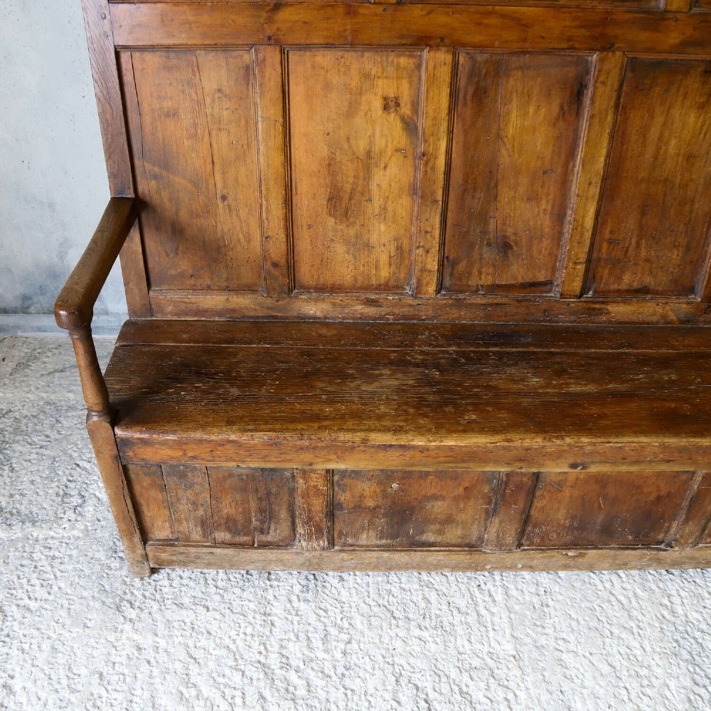 A high-backed antique wooden settle featuring raised timber panels, flat armrests with turned supports, and an enclosed panelled base. View VII