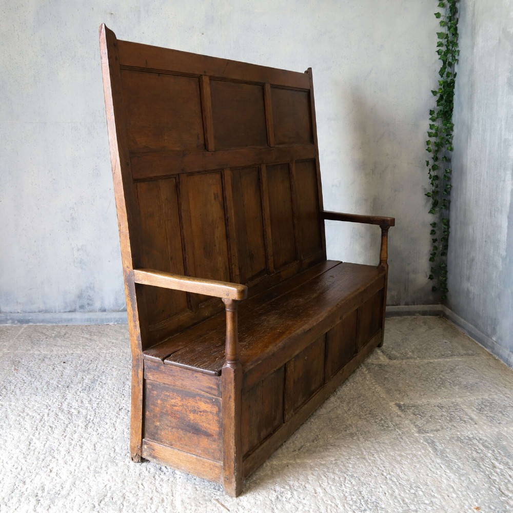 A high-backed antique wooden settle featuring raised timber panels, flat armrests with turned supports, and an enclosed panelled base. View XIV