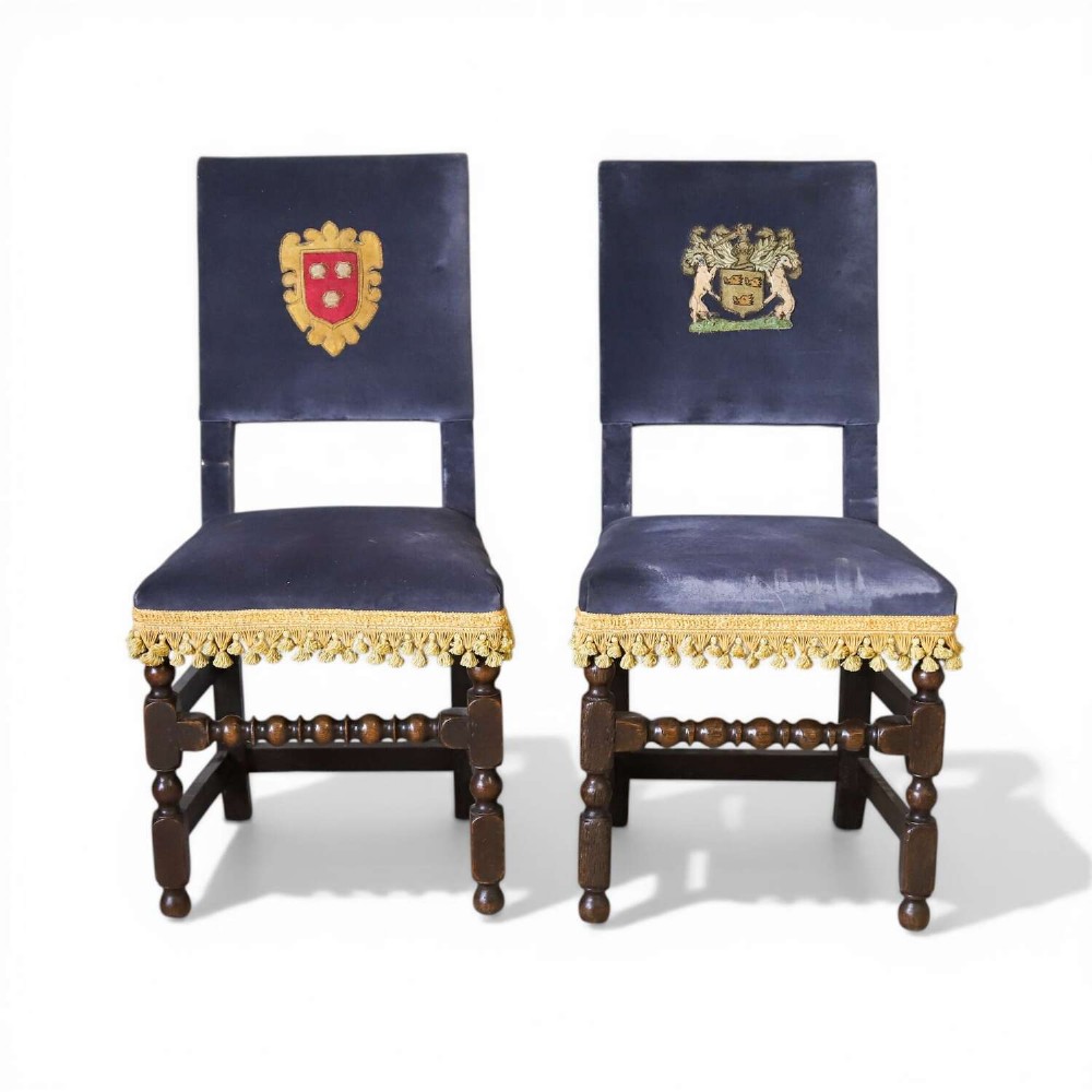 A matched pair of dark oak side chairs upholstered in dark blue velvet, featuring embroidered heraldic crests on the backrests and gold tassel fringe around the seat borders.