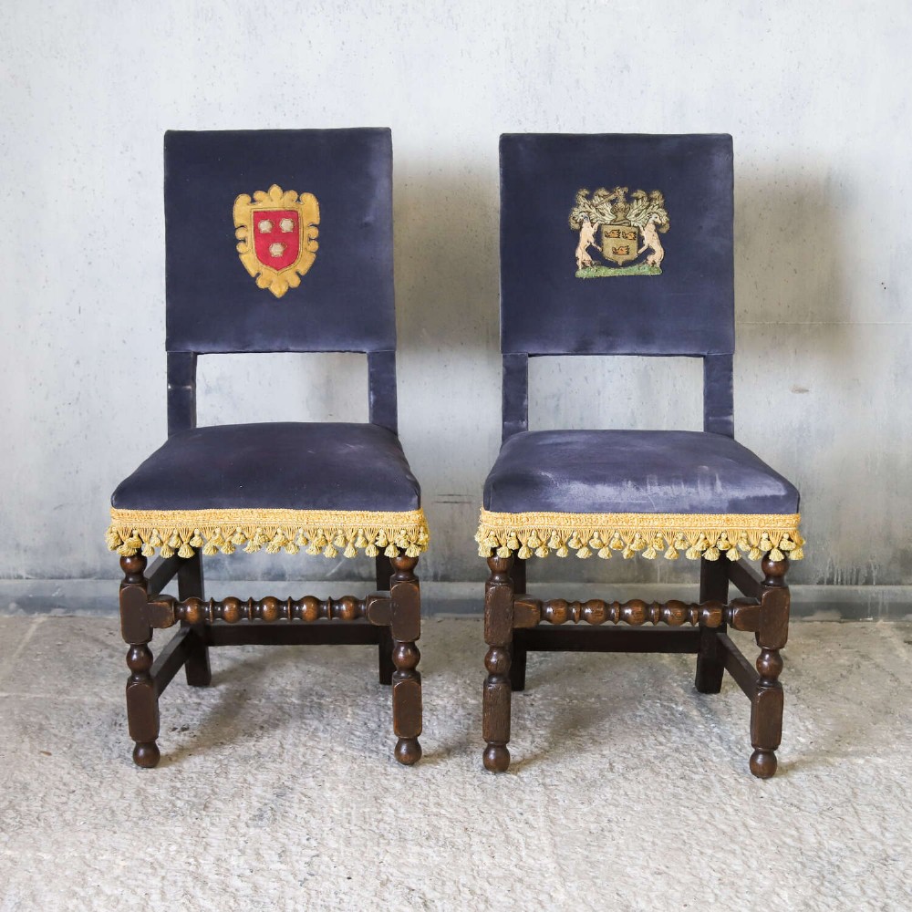 A matched pair of dark oak side chairs upholstered in dark blue velvet, featuring embroidered heraldic crests on the backrests and gold tassel fringe around the seat borders. View II
