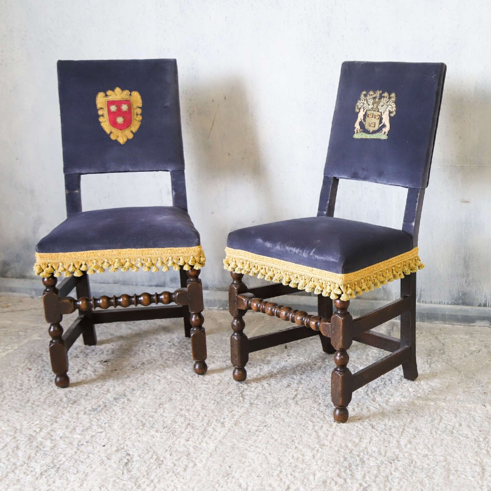 A matched pair of dark oak side chairs upholstered in dark blue velvet, featuring embroidered heraldic crests on the backrests and gold tassel fringe around the seat borders. View III