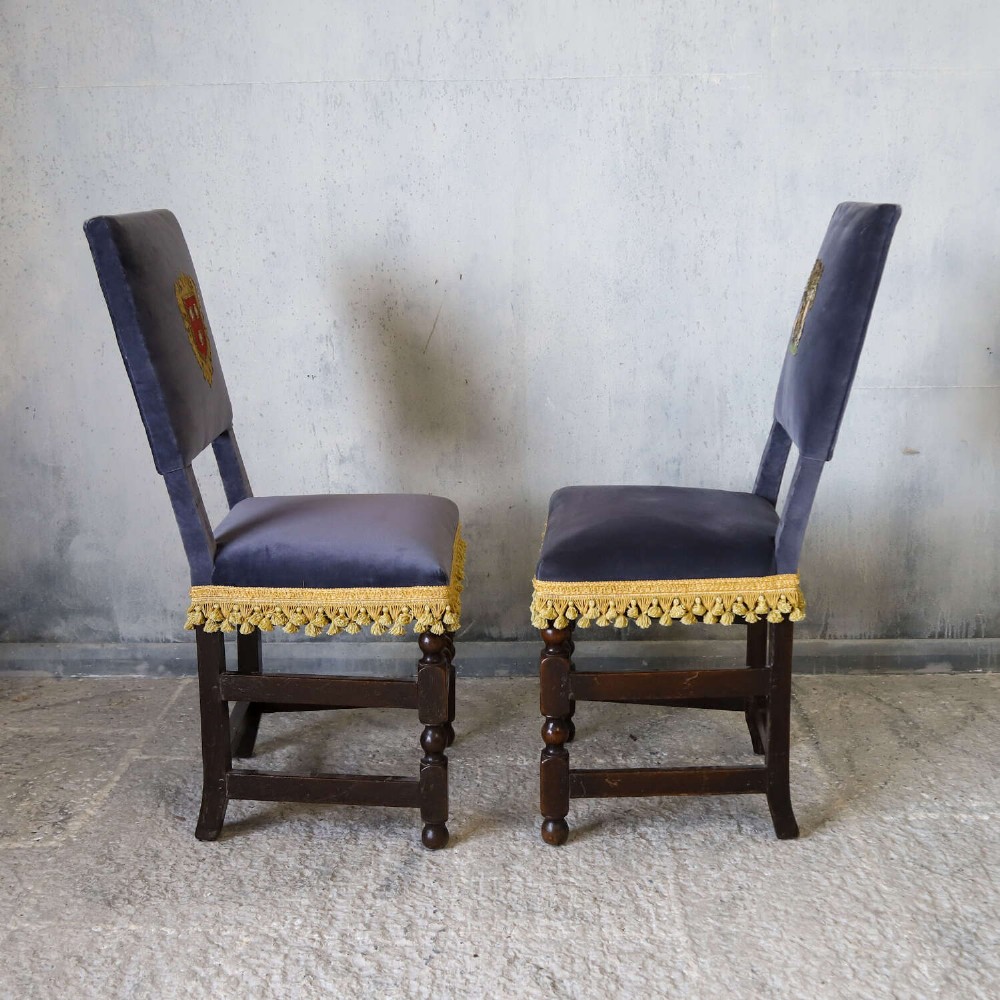 A matched pair of dark oak side chairs upholstered in dark blue velvet, featuring embroidered heraldic crests on the backrests and gold tassel fringe around the seat borders. View IX