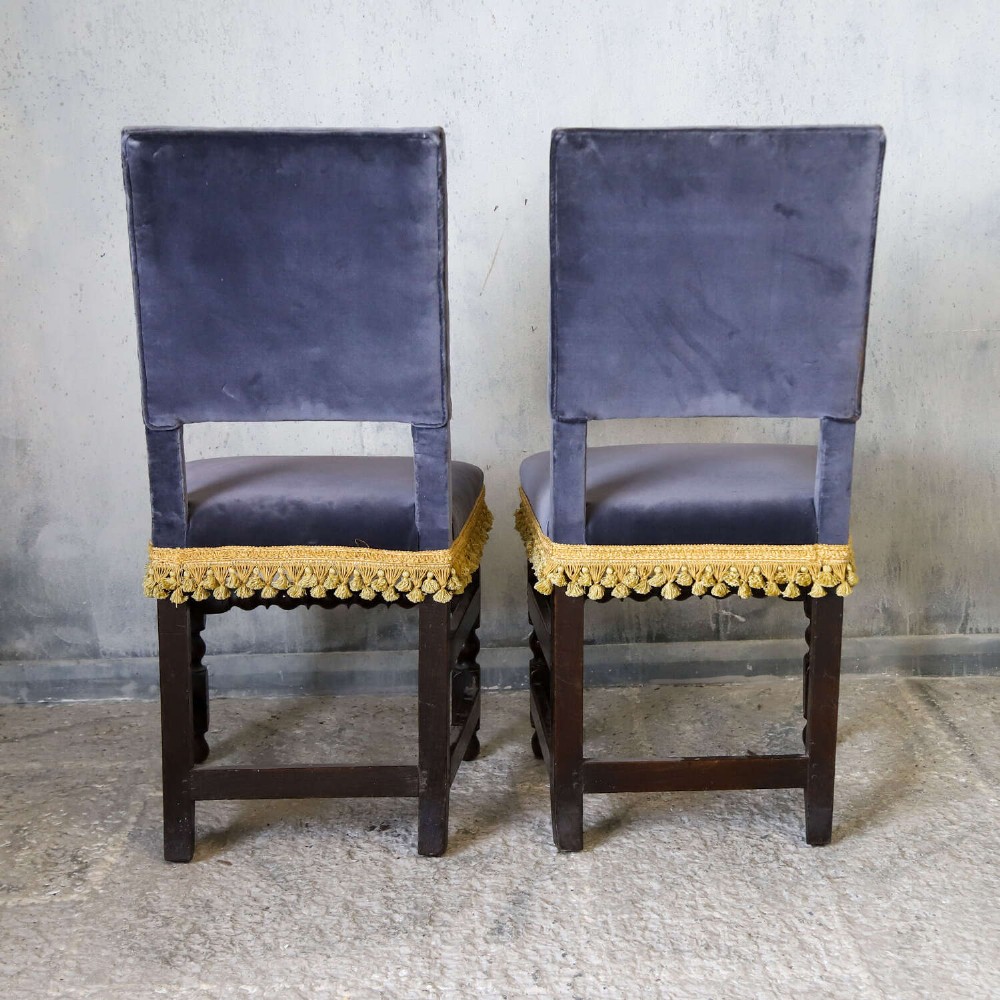 A matched pair of dark oak side chairs upholstered in dark blue velvet, featuring embroidered heraldic crests on the backrests and gold tassel fringe around the seat borders. View X