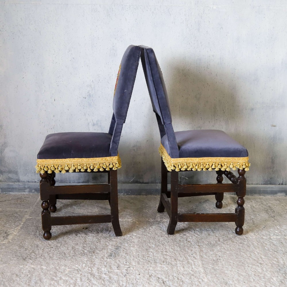 A matched pair of dark oak side chairs upholstered in dark blue velvet, featuring embroidered heraldic crests on the backrests and gold tassel fringe around the seat borders. View XI